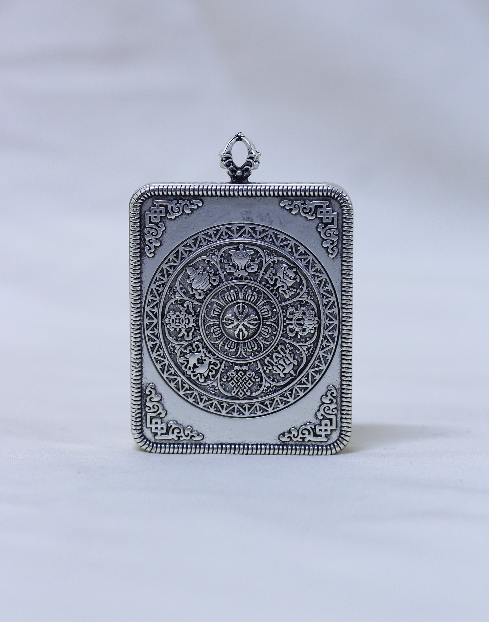 An ornate rectangular silver pendant featuring a detailed circular medallion and decorative border, photographed against a soft white background.