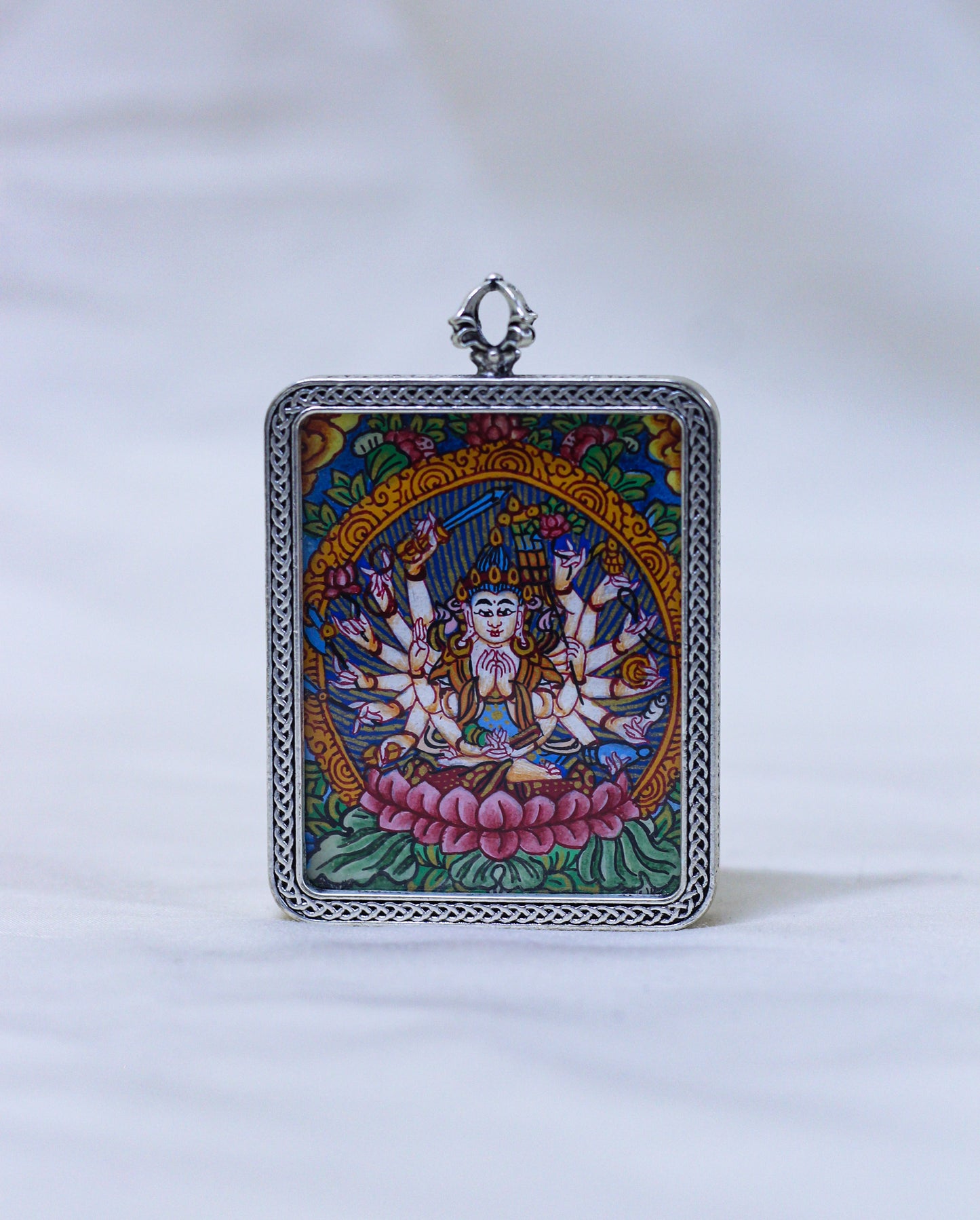 A square silver-toned pendant featuring a colorful Tibetan-style illustration of a multi-armed deity seated on a pink lotus, with a decorative silver border and top loop, shown against a white background.