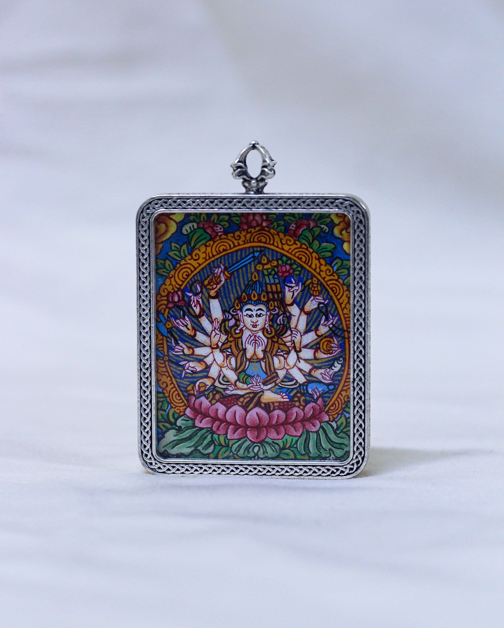 A square silver-toned pendant featuring a colorful Tibetan-style illustration of a multi-armed deity seated on a pink lotus, with a decorative silver border and top loop, shown against a white background.