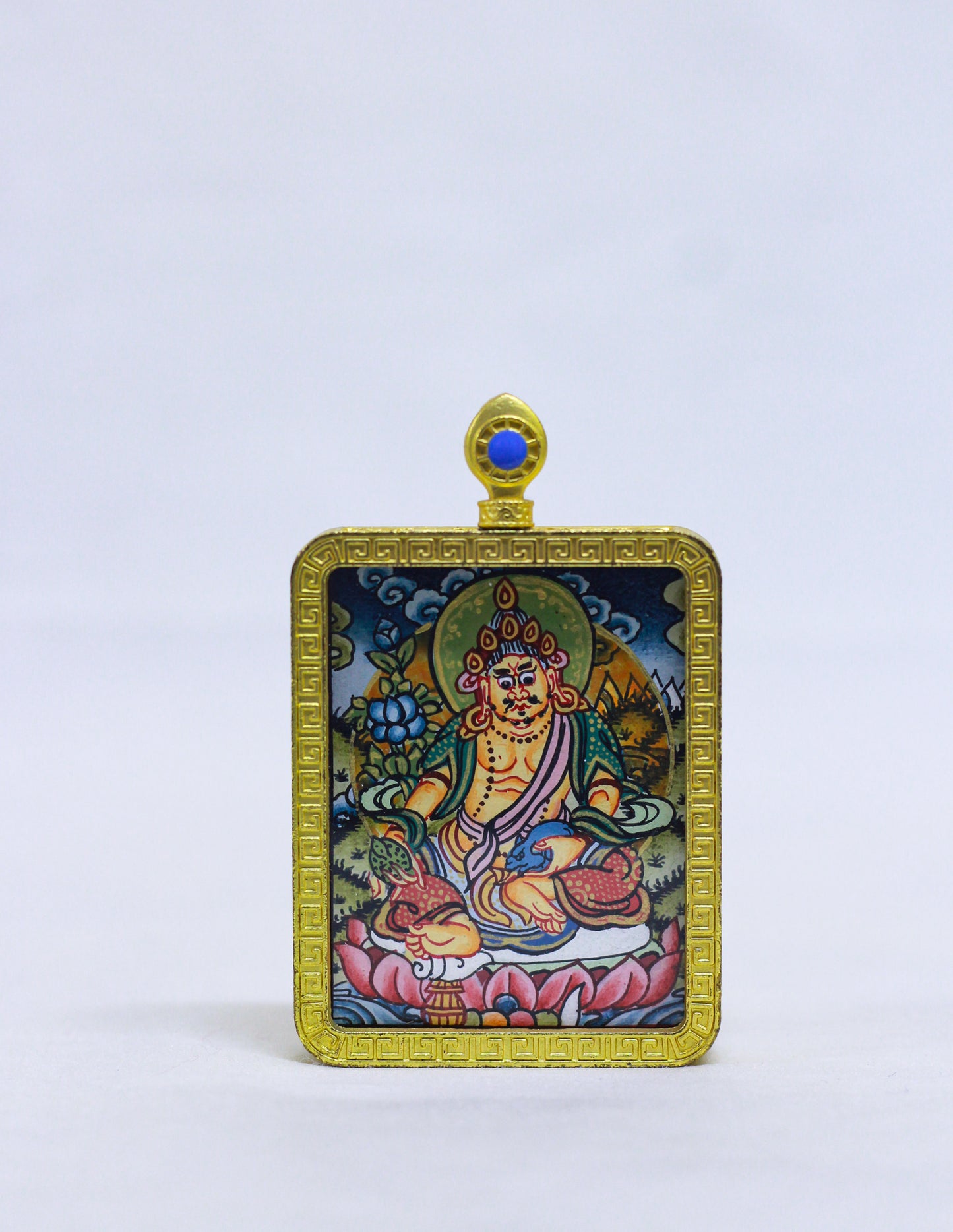 A rectangular Tibetan-style religious pendant or amulet featuring a Jambala illustration in a gold-toned frame, with a small blue stone at the top, photographed against a plain light background.