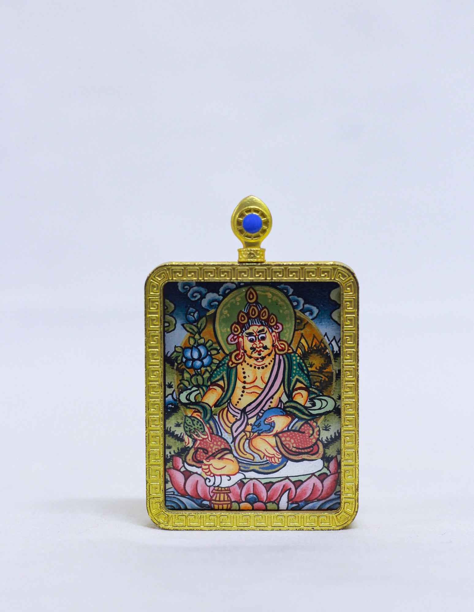 A rectangular Tibetan-style religious pendant or amulet featuring a Jambala illustration in a gold-toned frame, with a small blue stone at the top, photographed against a plain light background.