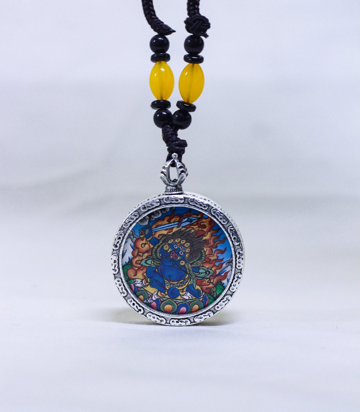 A circular silver-toned pendant necklace featuring a Mahakala thangka, suspended from a black cord with yellow and black beads.