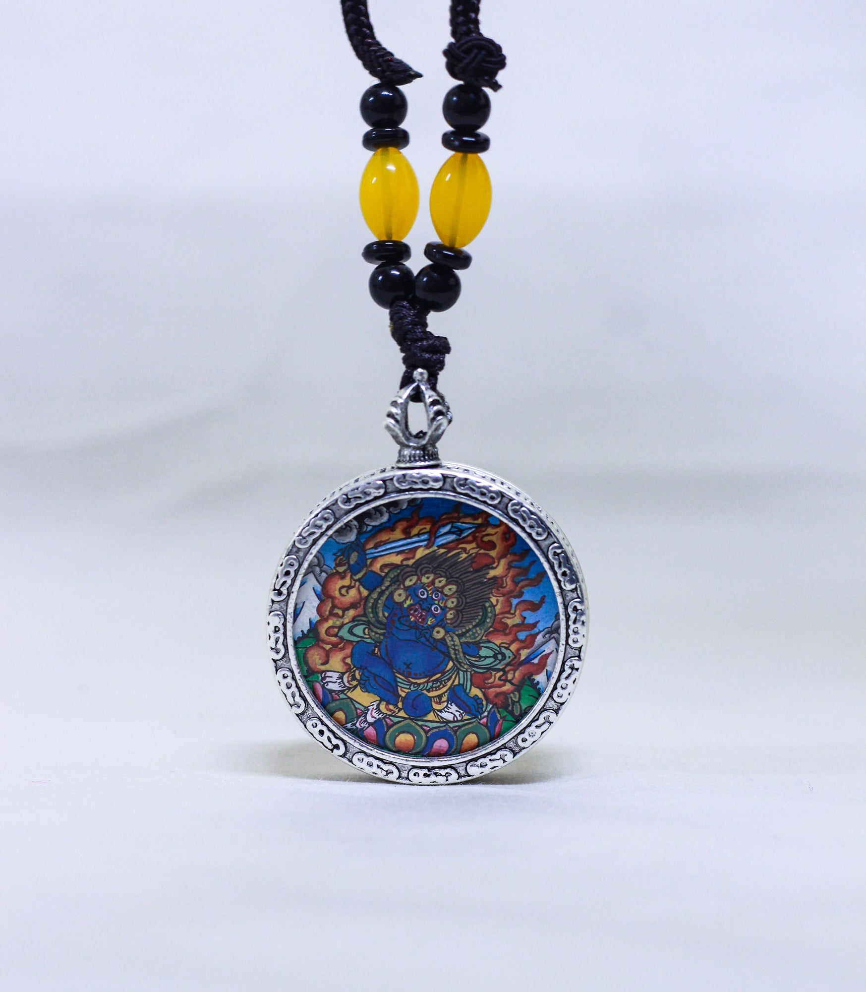 A circular silver-toned pendant necklace featuring a Mahakala thangka, suspended from a black cord with yellow and black beads.