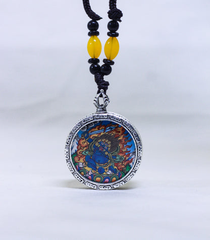 A circular silver-toned pendant necklace featuring a Mahakala thangka, suspended from a black cord with yellow and black beads.