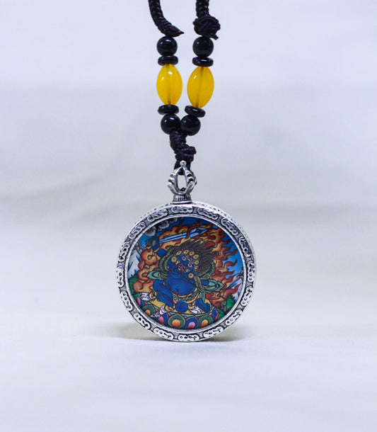 A circular silver-toned pendant necklace featuring a Mahakala thangka, suspended from a black cord with yellow and black beads.