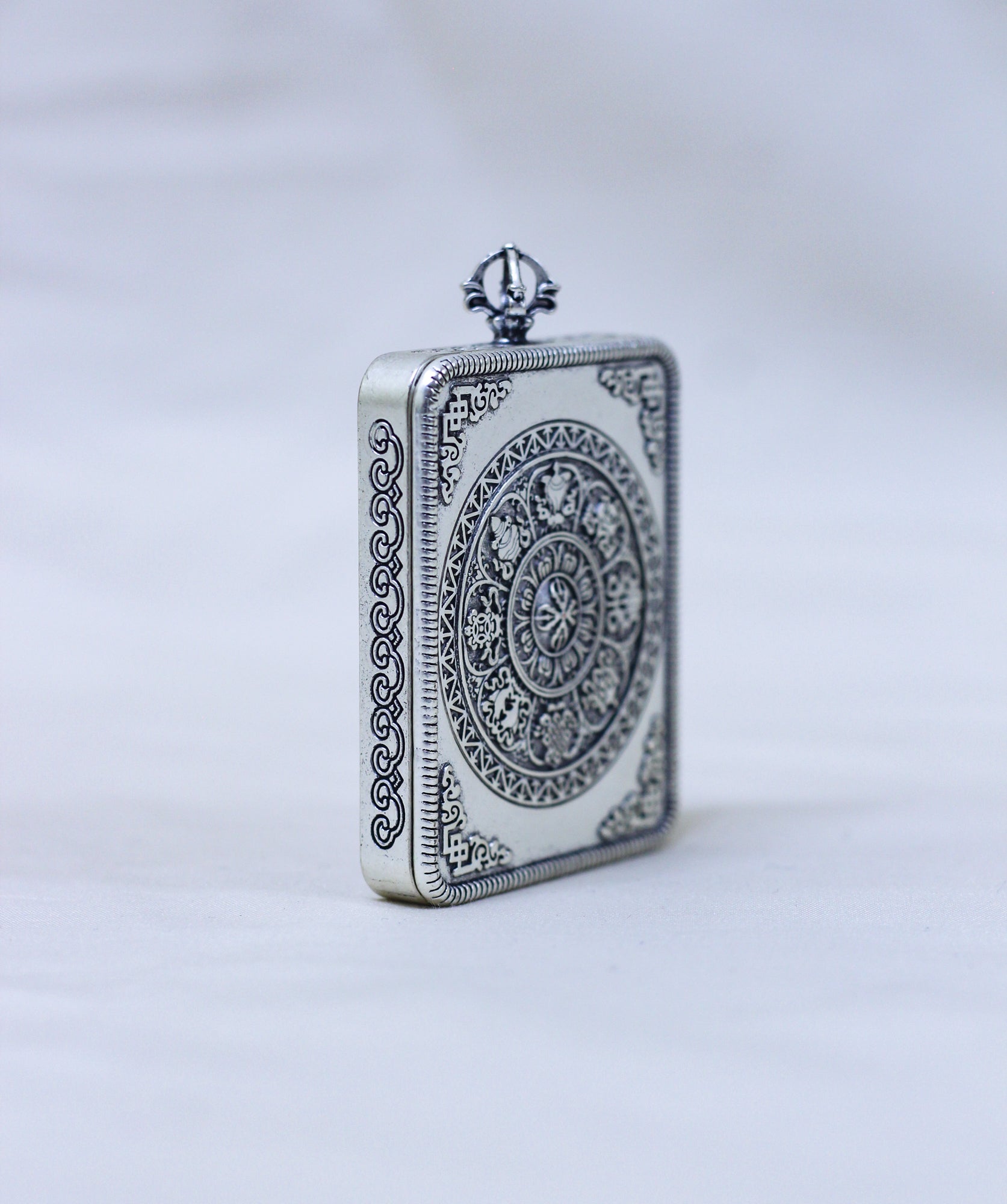 A rectangular silver-toned pendant or locket with ornate engraved circular and scroll patterns, standing upright on a light gray background.