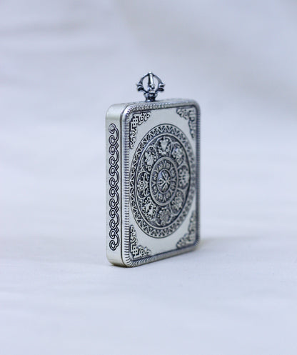 A rectangular silver-toned pendant or locket with ornate engraved circular and scroll patterns, standing upright on a light gray background.