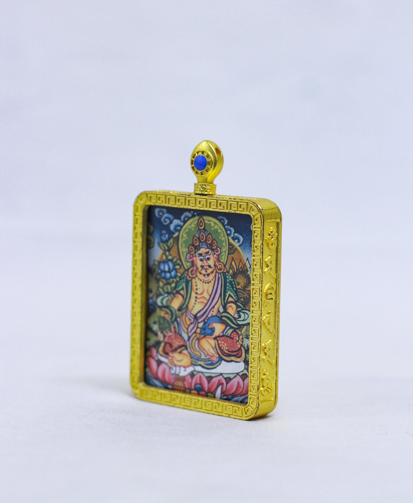 Side view of a small rectangular gold-toned pendant with a religious illustration of Jambala in the center and a blue stone detail at the top where there is a hoop.