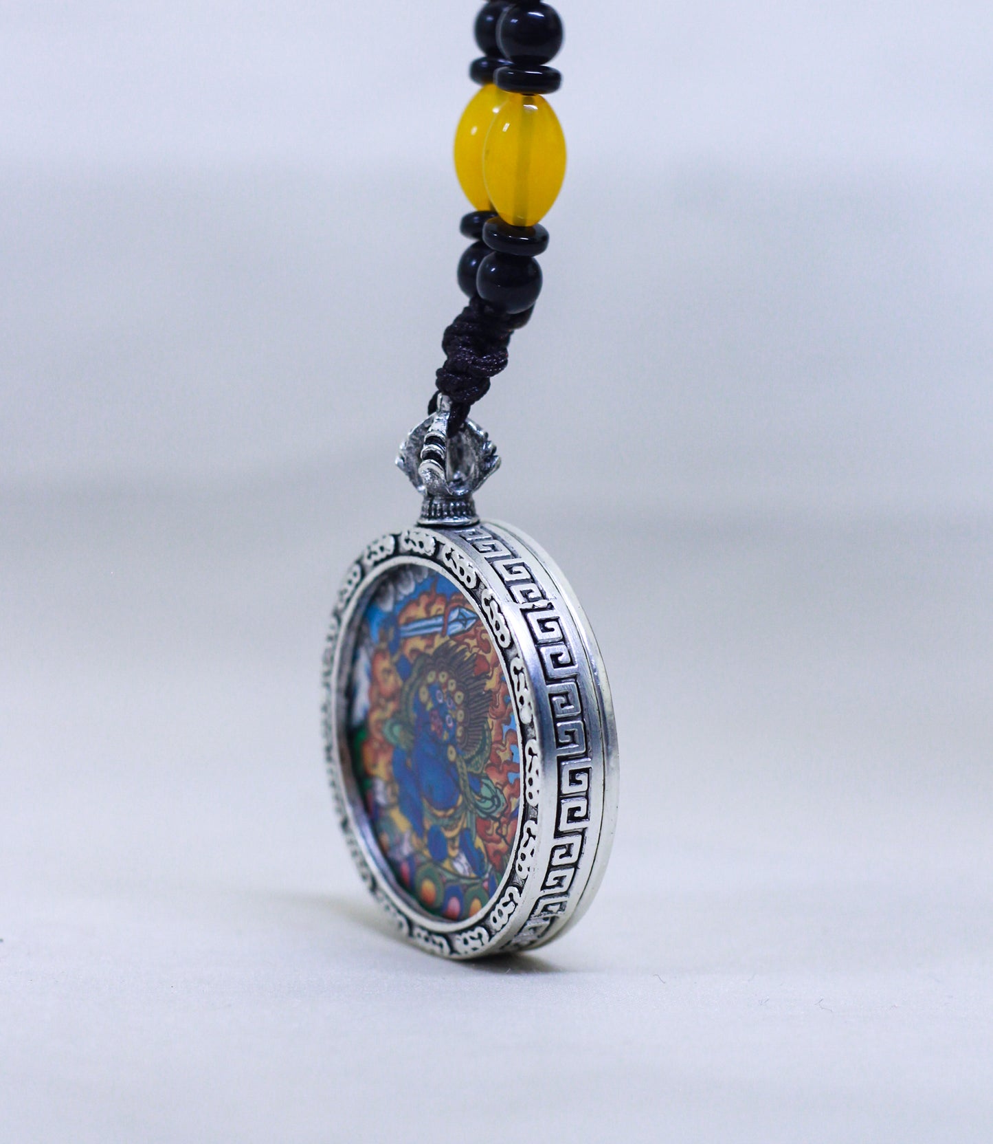 A side view of a silver-toned circular Mahakala butti thangka pendant featuring a colorful bead, suspended from a black cord with a yellow accent bead.
