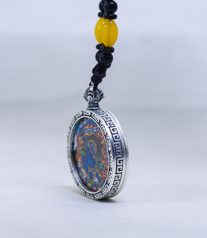 A side view of a silver-toned circular Mahakala butti thangka pendant featuring a colorful bead, suspended from a black cord with a yellow accent bead.