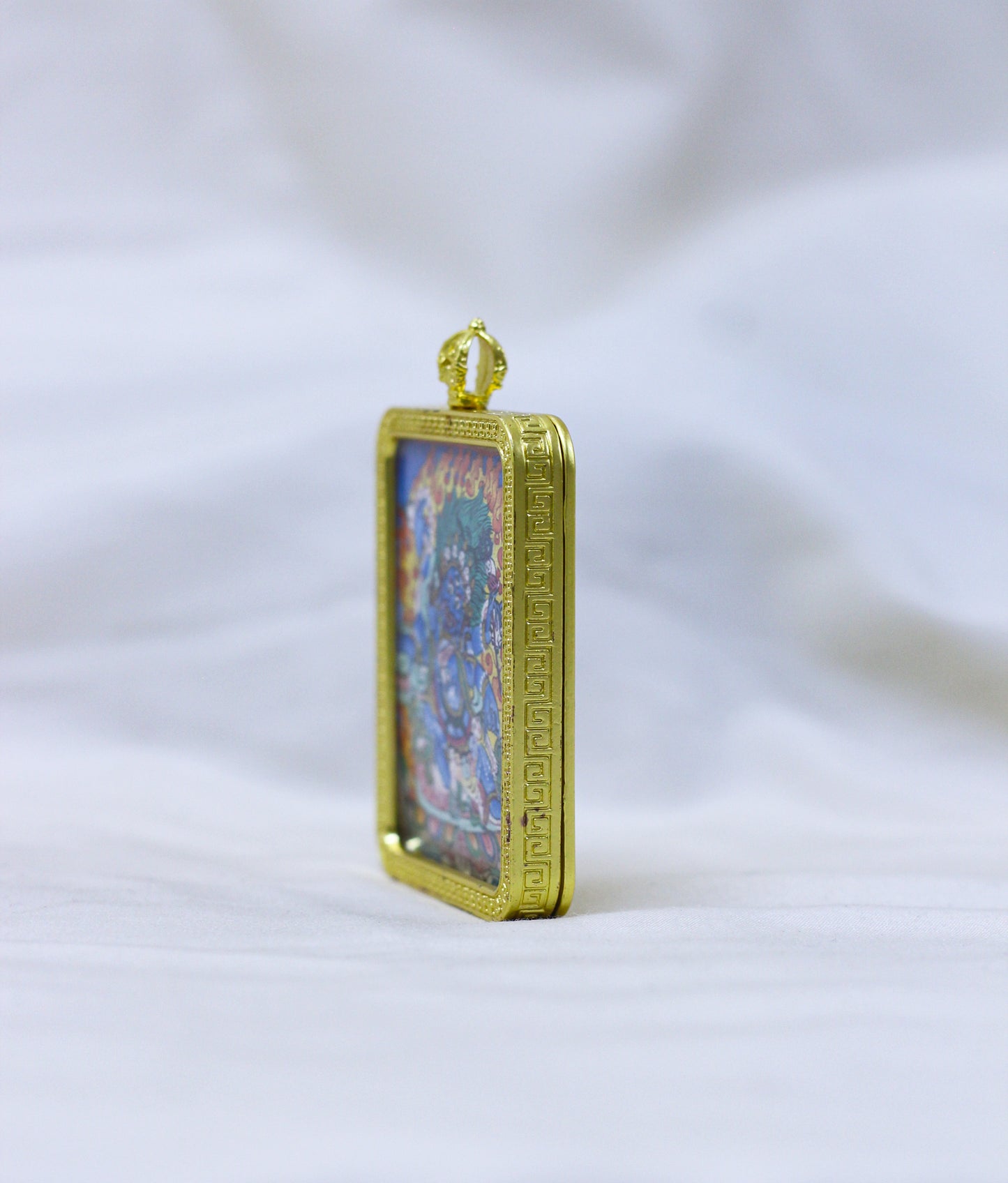 Side view of the Mahakala thangka pendant, a gold-toned border pendant with an embossed border standing upright 
