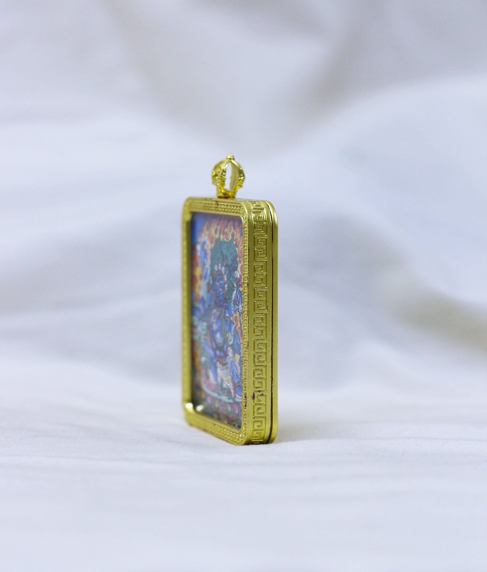 Side view of the Mahakala thangka pendant, a gold-toned border pendant with an embossed border standing upright 
